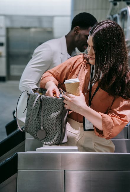 A woman with long dark hair, wearing an orange-brown jacket, is standing at an airport baggage claim area, reaching into a grey fabric handbag with circular branding, to retrieve a passport and a small paper cup she is holding in her right hand. Behind her, a man with short black hair, dressed in a white shirt, is also at the baggage carousel, appearing to assist or wait. The scene is set indoors with metallic and glass surfaces, and the lighting is bright and neutral. The image captures a moment of home relocation or travel, emphasizing packing and moving logistics relevant to house removals or transport services, such as those provided by Removal Companies Islington.