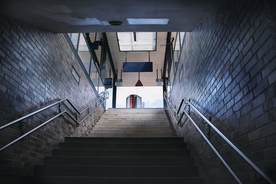 View of the staircase leading from an underground passage at Angel station, Islington, with brick walls on either side and metal handrails. The staircase is illuminated by natural daylight from the open upper entrance, revealing a glimpse of the platform signage and a ticket machine in the background. This scene depicts an interior setting relevant to house removals and luggage transport, as part of the typical building access points involved in home relocation services offered by Removal Companies Islington. The image emphasizes the structural details of the stairs, the brickwork, and the transition from underground to street level, supporting the process of packing, loading, or moving furniture and boxes through single or multiple levels during a building access process.