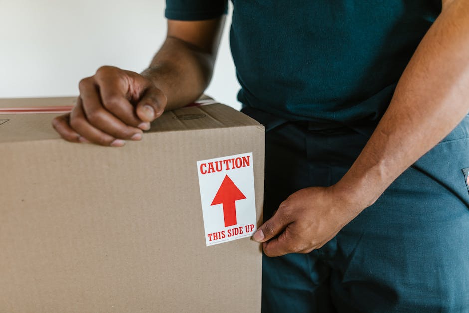 A person wearing a dark green shirt and navy blue trousers is holding a large cardboard box during a home relocation process, inside an indoor space near a doorway with a tiled floor. The person is gr