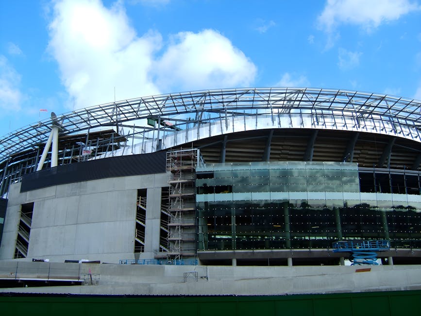 A modern building under construction with a curved steel framework at the top, partly covered with glass panels and scaffolding on the side. The structure is situated on a concrete base, with an open area at ground level. The sky above is bright blue with some scattered white clouds, and construction materials and equipment are visible around the site. This image relates to the process of home relocation and furniture transport, illustrating the logistical aspects of moving large fixtures during building construction or renovation, which may be relevant to house removals and packing and moving services offered by Removal Companies Islington.