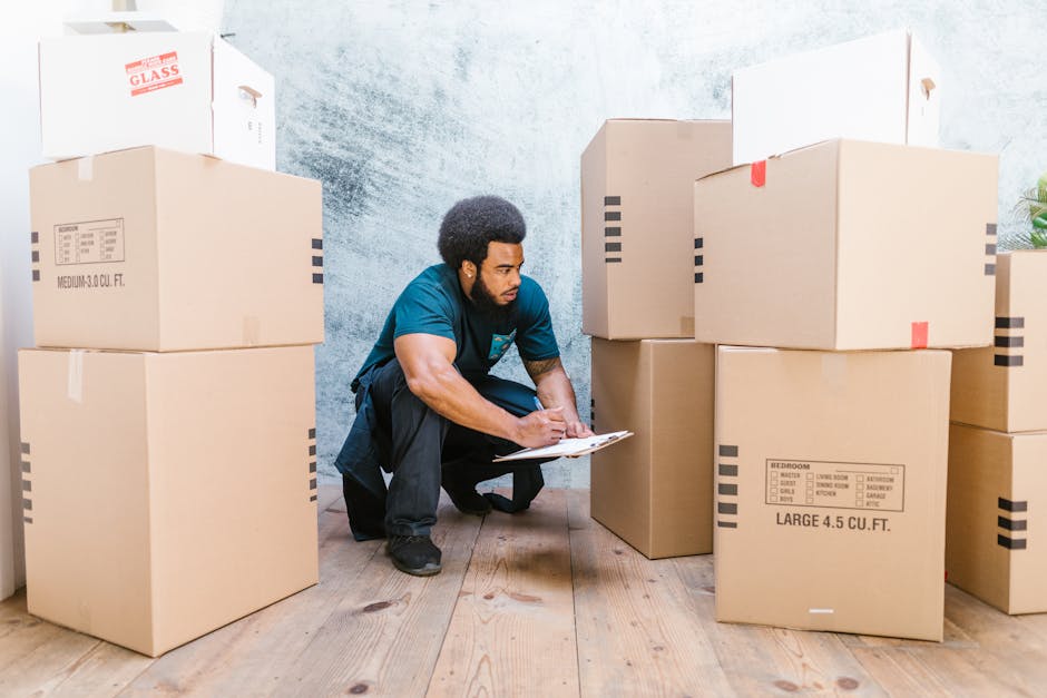 A man crouching on a wooden floor inside a property, surrounded by multiple large cardboard moving boxes, some labeled with room contents and sizes, with a clipboard in hand, appearing to check or record inventory during a home relocation process. The boxes are stacked and arranged around him, with additional boxes on a nearby window sill and a doorway visible in the background. The scene illustrates packing and moving activities as part of a furniture transport or home removals procedure, consistent with services offered by Removal Companies Islington. The lighting is natural, and the environment suggests an interior space in the midst of a professional move, highlighting the careful organization and preparatory steps involved in relocation and packing services.
