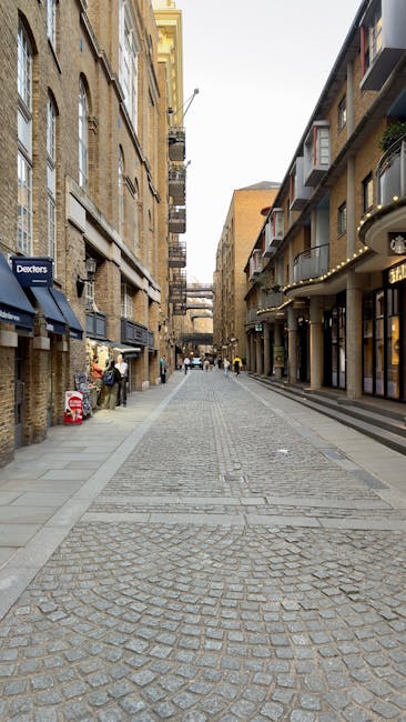 A wide view of a narrow urban street in Islington with cobblestone paving, flanked by multi-storey buildings with a mix of brickwork and modern façades. The street is lined with commercial premises on the left, including a shop with a blue awning, and residential or commercial buildings on the right with balconies and large windows. There are a few pedestrians further down the street. In the foreground, a small moving truck or van, associated with Removal Companies Islington, is parked with its rear doors open, part of the loading process visible. Packing materials such as cardboard boxes and plastic wrap are visible inside the vehicle, ready for a home relocation or furniture transport. Some items, including a piece of furniture wrapped in fabric and packing boxes, are being carried or loaded into the van, with a trolley and straps visible on the pavement nearby, indicating an active luggage and furniture moving operation. The scene captures the typical urban environment during a residential move involving careful packing, loading, and transportation within the narrow lanes of Camden Passage in Islington. The lighting is natural, suggesting daytime, with shadows cast on the cobbled street, reflecting a professional and organized approach to house removals.