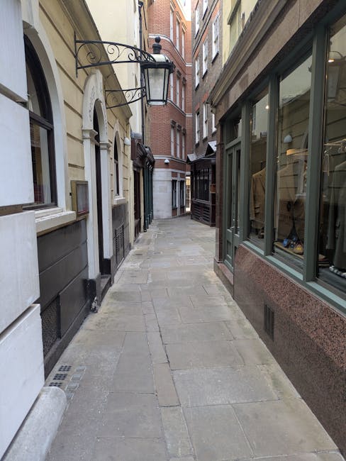 A wide view of a narrow urban street in Islington with cobblestone paving, flanked by multi-storey buildings with a mix of brickwork and modern façades. The street is lined with commercial premises on the left, including a shop with a blue awning, and residential or commercial buildings on the right with balconies and large windows. There are a few pedestrians further down the street. In the foreground, a small moving truck or van, associated with Removal Companies Islington, is parked with its rear doors open, part of the loading process visible. Packing materials such as cardboard boxes and plastic wrap are visible inside the vehicle, ready for a home relocation or furniture transport. Some items, including a piece of furniture wrapped in fabric and packing boxes, are being carried or loaded into the van, with a trolley and straps visible on the pavement nearby, indicating an active luggage and furniture moving operation. The scene captures the typical urban environment during a residential move involving careful packing, loading, and transportation within the narrow lanes of Camden Passage in Islington. The lighting is natural, suggesting daytime, with shadows cast on the cobbled street, reflecting a professional and organized approach to house removals.