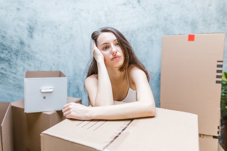 A young woman with long brown hair, wearing a white tank top, is resting her head on her left hand while sitting behind a large cardboard box. Around her are several packing boxes made of brown cardboard, some closed and others open, placed in what appears to be a home or storage area with a textured gray wall in the background. The scene suggests she is in the process of packing or unpacking during a home relocation. Visible in the scene are packing materials such as cardboard boxes, and the setting indicates a moving or furniture transport context. This image reflects the logistical and emotional aspects often associated with house removals, which companies like Removal Companies Islington manage as part of their removals services, including packing, loading, and transporting household items.
