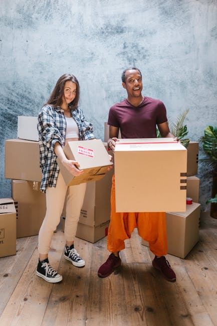 Two individuals engaged in a home relocation process, standing indoors against a textured, light blue wall with a wooden floor. The person on the left, a young woman with long brown hair, is dressed casually in a checkered shirt, beige pants, and sneakers, holding a cardboard box with a red and white 'Fragile' label. The person on the right, a man with short hair and a dark complexion, is wearing a maroon t-shirt and orange trousers, holding multiple cardboard boxes of varying sizes, some wrapped in plastic or padding materials for protection during furniture transport. Several packed boxes and moving supplies are arranged around them, indicating an active packing and loading process typical of house removals. Visible in the environment are also plastic wrapping and packing tape, supporting the context of packing and moving services provided by companies such as Removal Companies Islington, specializing in furniture transport and home relocation.