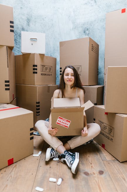 A young woman with long brown hair, wearing a white tank top, is resting her head on her left hand while sitting behind a large cardboard box. Around her are several packing boxes made of brown cardboard, some closed and others open, placed in what appears to be a home or storage area with a textured gray wall in the background. The scene suggests she is in the process of packing or unpacking during a home relocation. Visible in the scene are packing materials such as cardboard boxes, and the setting indicates a moving or furniture transport context. This image reflects the logistical and emotional aspects often associated with house removals, which companies like Removal Companies Islington manage as part of their removals services, including packing, loading, and transporting household items.