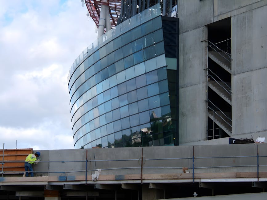 A construction worker wearing a high-visibility yellow jacket and helmet is standing on a scaffolded platform outside a modern building with a curved glass facade, reflecting the sky and nearby structures. To the right, the building features a concrete exterior with an accessibility stairwell enclosed by metal railings. The scene is set on a pavement adjacent to the building, with some construction materials and equipment visible, including a large wooden formwork panel and tools. The image captures part of the ongoing construction or renovation process, illustrating the typical environment for house removals or building refurbishment projects that Removal Companies Islington may handle, particularly emphasizing loading or relocation logistics on-site.