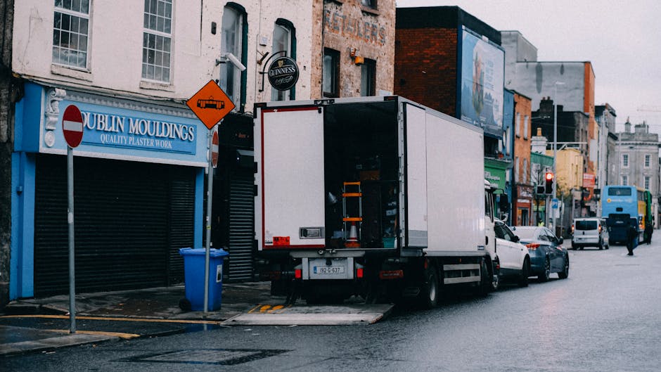 A white moving truck with its rear door open is parked on a street in Islington, adjacent to a storefront with a blue sign reading 'Dublin Mouldings' offering plaster work. The truck is positioned next to a sidewalk, with the loading area extended onto the street for furniture transport and packing materials. Inside the truck, visible packing supplies such as cardboard boxes and wrapping materials are prepared for home relocation. A blue wheeled trolley and lifting straps are used to assist with the loading process. Several cardboard boxes and smaller items are being carried towards the truck, with some placed nearby on the pavement. Behind the truck, other vehicles, including white cars and a blue bus, are seen driving along the street, while commercial buildings and signage line the background. The scene depicts a typical urban environment for a residential move, emphasizing the logistics involved in packing and loading for a house removal, with the presence of secure packaging materials and equipment representing professional moving services by Removal Companies Islington.