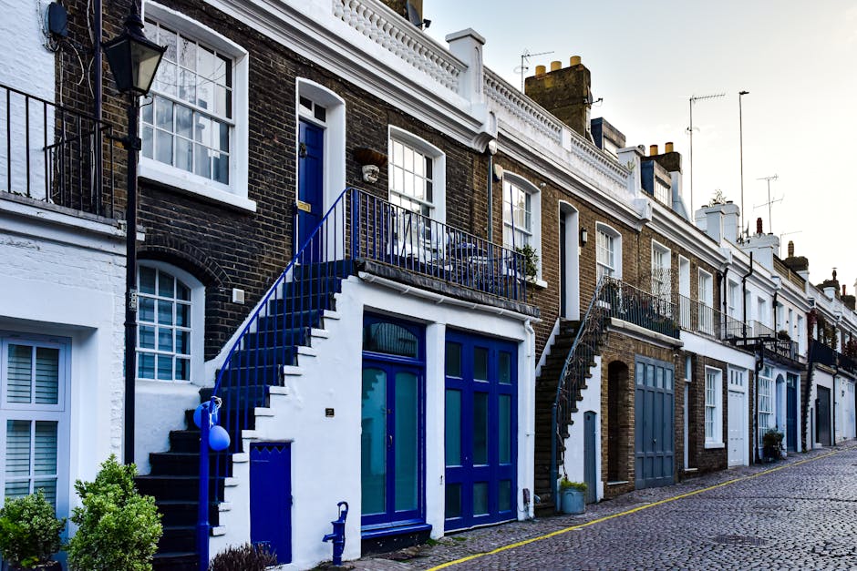 A row of terraced houses in Islington with brick facades and white window frames, featuring small front gardens and balcony railings. The houses have external staircases leading from the pavement to upper-level entrances, with one staircase painted in a striking blue color. Some houses have large ground-floor windows and glass doors, indicating possible main entrances or assistance with home relocation. The street is paved with cobblestones, and the scene is lit by natural daylight, suggesting a daytime moving or packing process. A large van or transport vehicle could be nearby but is not visible in the image. The presence of boxes and furniture is not shown, but the scene reflects typical urban properties where careful furniture transport and packing may be necessary, as handled by companies like Removal Companies Islington during their house removals or moving services.