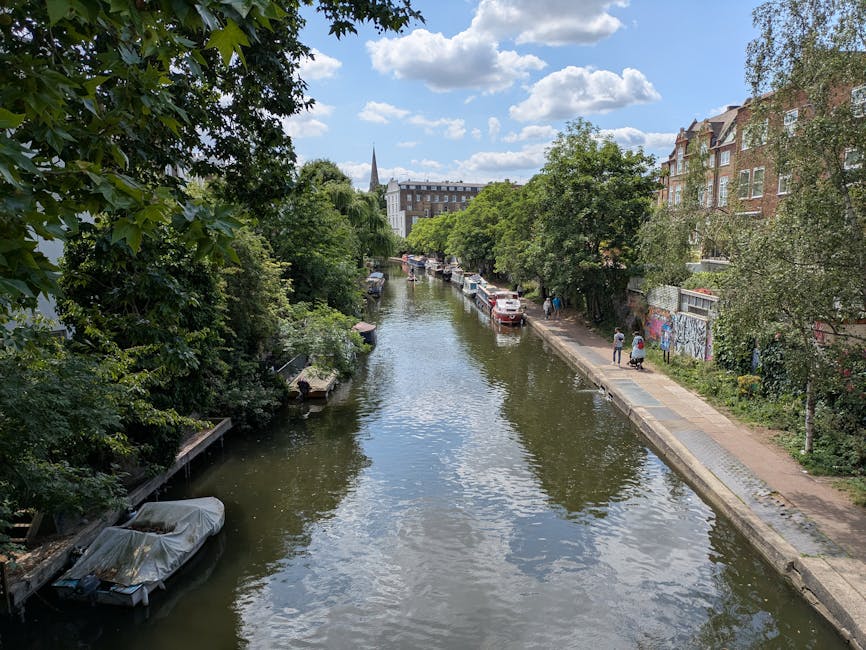 A scenic view of a narrow canal lined with lush green trees and residential buildings in Islington during daylight. Several parked cars are visible along the canal-side pavement, with a few people walking on the path. A small boat, covered with a protective tarp, is moored on the left side of the canal. In the background, a church steeple rises above the trees, and the sky is partly cloudy with patches of blue. The image captures the peaceful atmosphere typical of an urban home relocation area, with the environment suitable for moving logistics and transport activities supported by Removal Companies Islington.