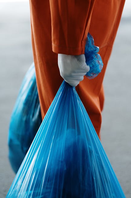 Close-up view of a person wearing an orange jumpsuit and white gloves, holding a blue plastic bag filled with waste or debris, during a home relocation or clearance process. The individual is standing outdoors on a paved surface, with another similar blue bag placed on the ground nearby. The scene suggests the handling of waste materials as part of a residential move, possibly involving clearing or disposing of bulky waste items in compliance with Islington disposal rules. The lighting is natural, and the focus is on the manual effort involved in lifting and transporting waste, emphasizing the logistical aspects of packing and moving tasks undertaken by Removal Companies Islington in the context of property relocation services.