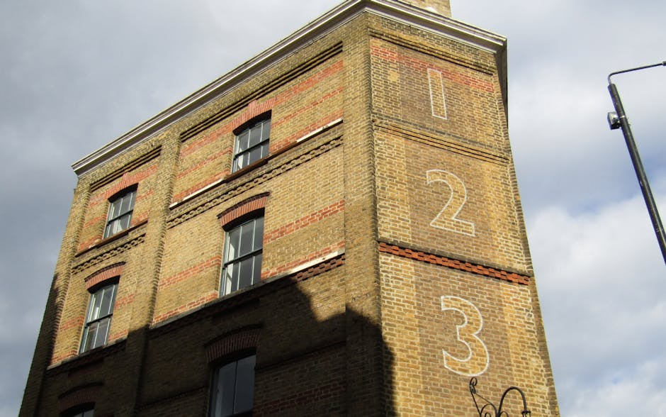 Photograph of a multi-storey brick building with three visible windows, each fitted with black frames and white curtains. The building's facade features a mix of tan and reddish-brown bricks, with red brick arches above the windows. Large, white painted numbers '1', '2', and '3' are vertically aligned on the right side of the building, indicating the floors. The photograph is taken from a low angle, capturing the upper floors and part of the sky, which is partly cloudy with grey and white clouds. A street lamp is partially visible on the right edge of the image. The scene suggests an urban environment consistent with house and apartment block renovations or relocations, and could be relevant to house removal or moving logistics in the Islington area, as managed by Removal Companies Islington.