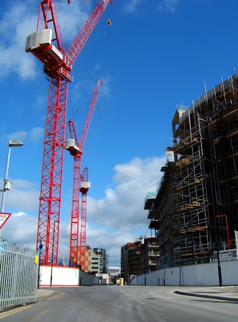 A construction site on a clear day with a bright blue sky, featuring two tall red tower cranes with white operator cabins, lifting equipment, and long booms extending overhead. To the right, there is a partially completed multi-storey building surrounded by scaffolding, with exterior floors lined with safety barriers and netting. The site is enclosed by a metal fence with white and red panels at the base, and the foreground shows a paved street with a street lamp and some road signage. This environment illustrates ongoing building development, relevant to busy urban environments where house removals and home relocation services, such as those offered by Removal Companies Islington, may operate, especially when coordinating moving logistics around construction areas.
